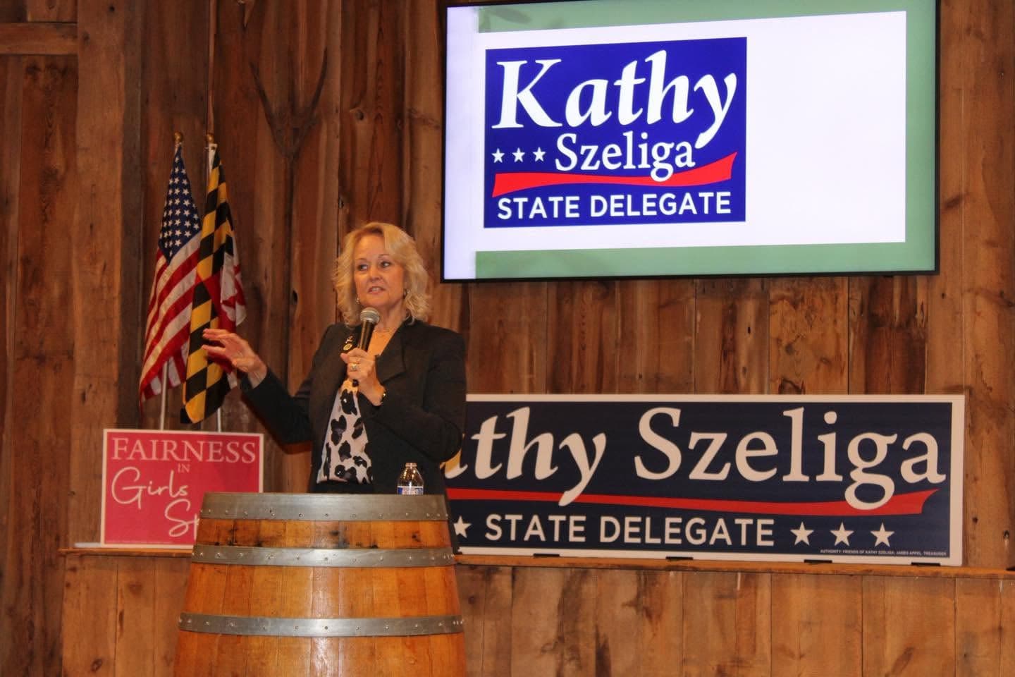 Kathy Szeliga speaking at a wine-barrel podium inside the Boordy Vineyards barn, flags and campaign signs behind her.