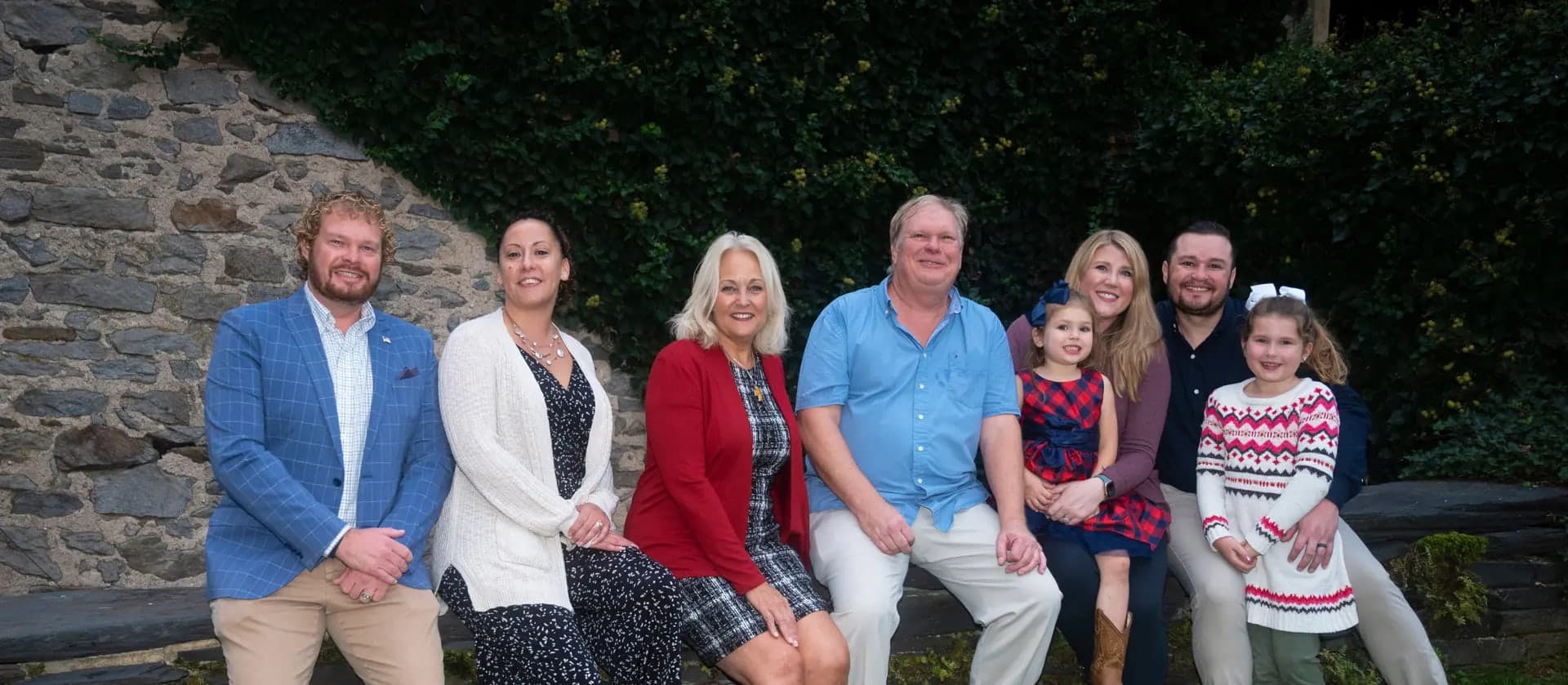 Kathy and Mark Szeliga seated on a stone wall with their sons, daughters-in-law, and two granddaughters.