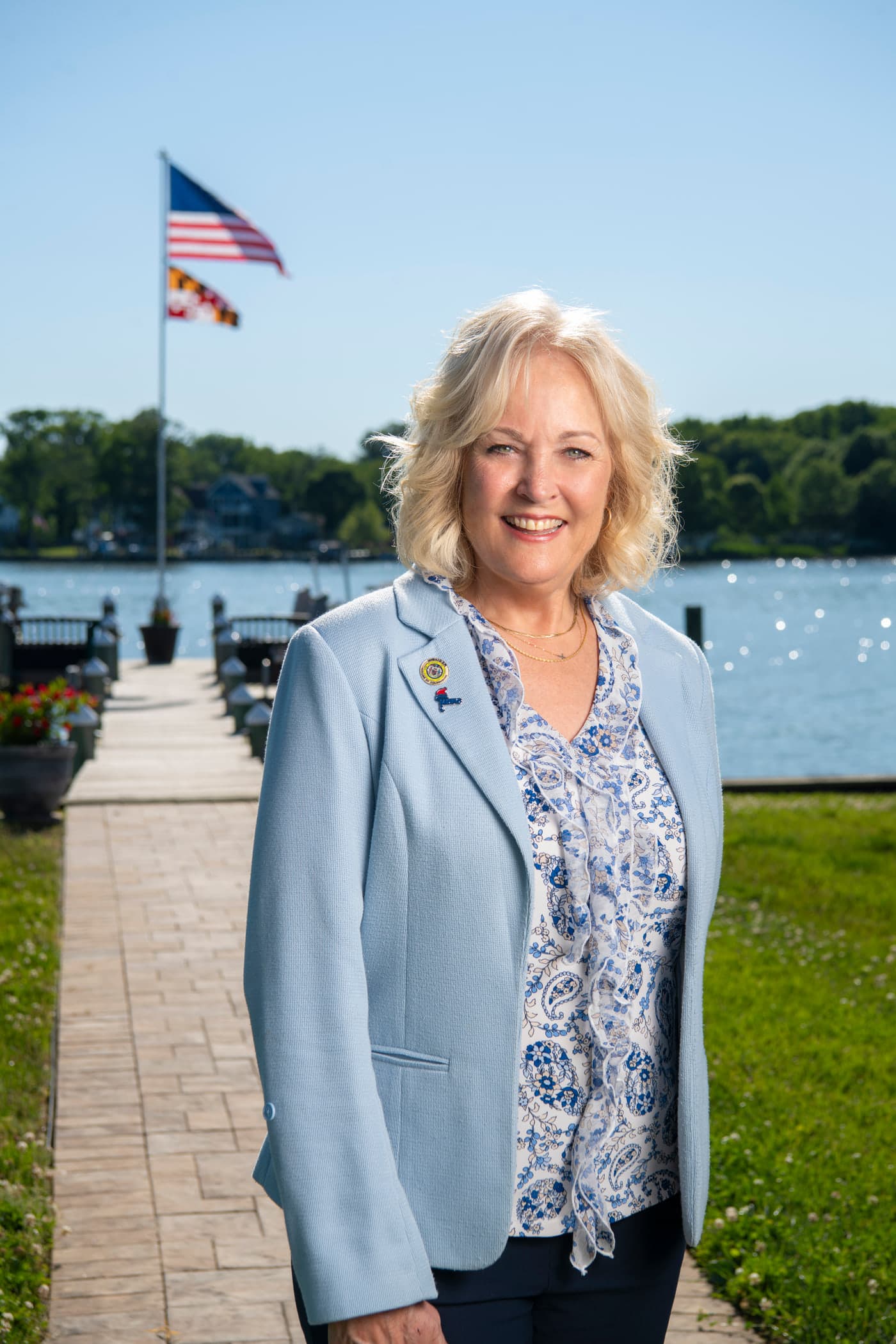 Delegate Kathy Szeliga in a light blue blazer on a waterfront promenade, the Maryland flag flying behind her.
